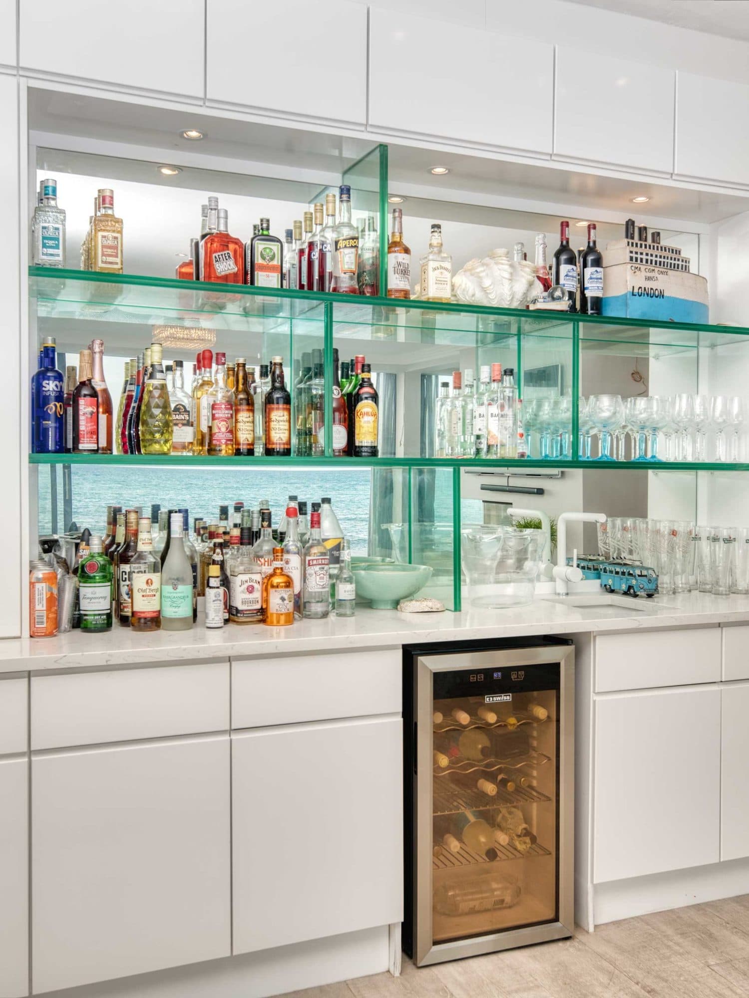 A fully stocked glass-shelved bar with the ocean visible through the window behind it